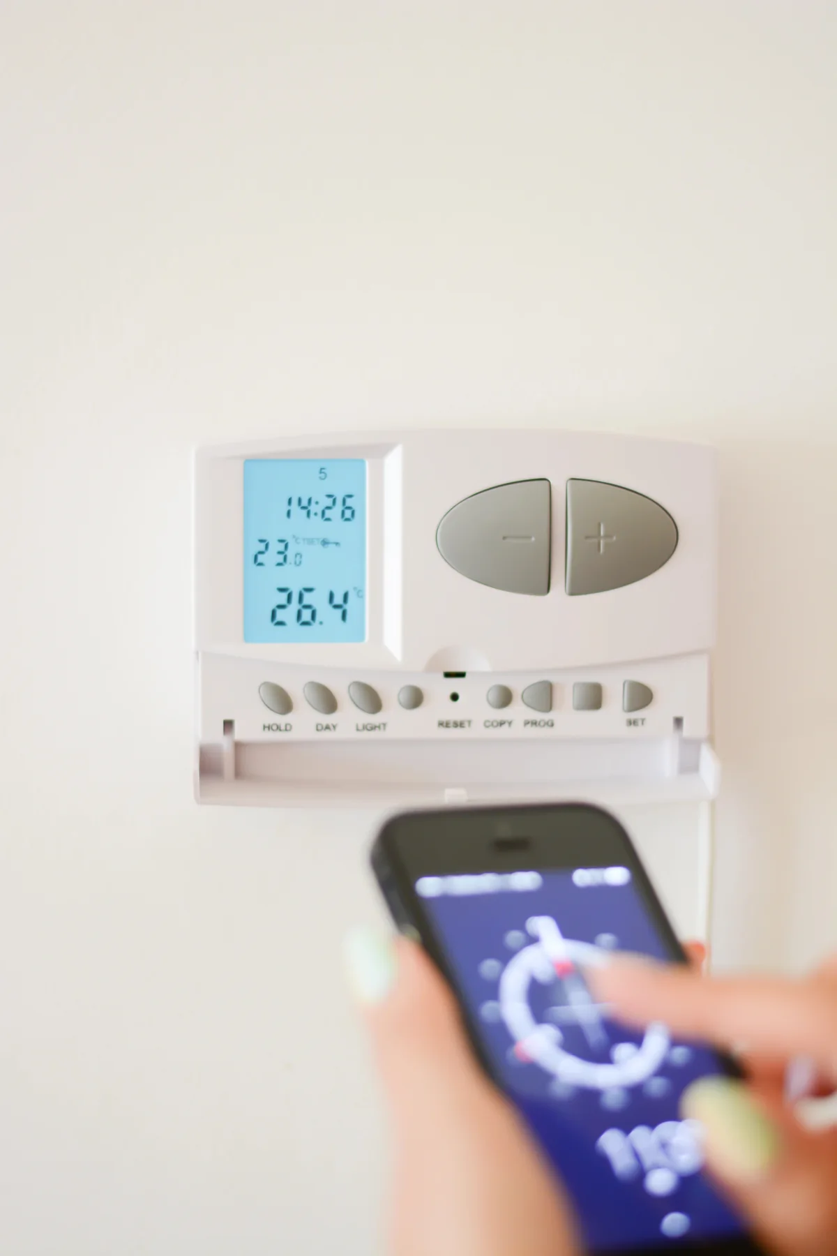 Heating controls shown on a mobile phone with the wall thermostat behind